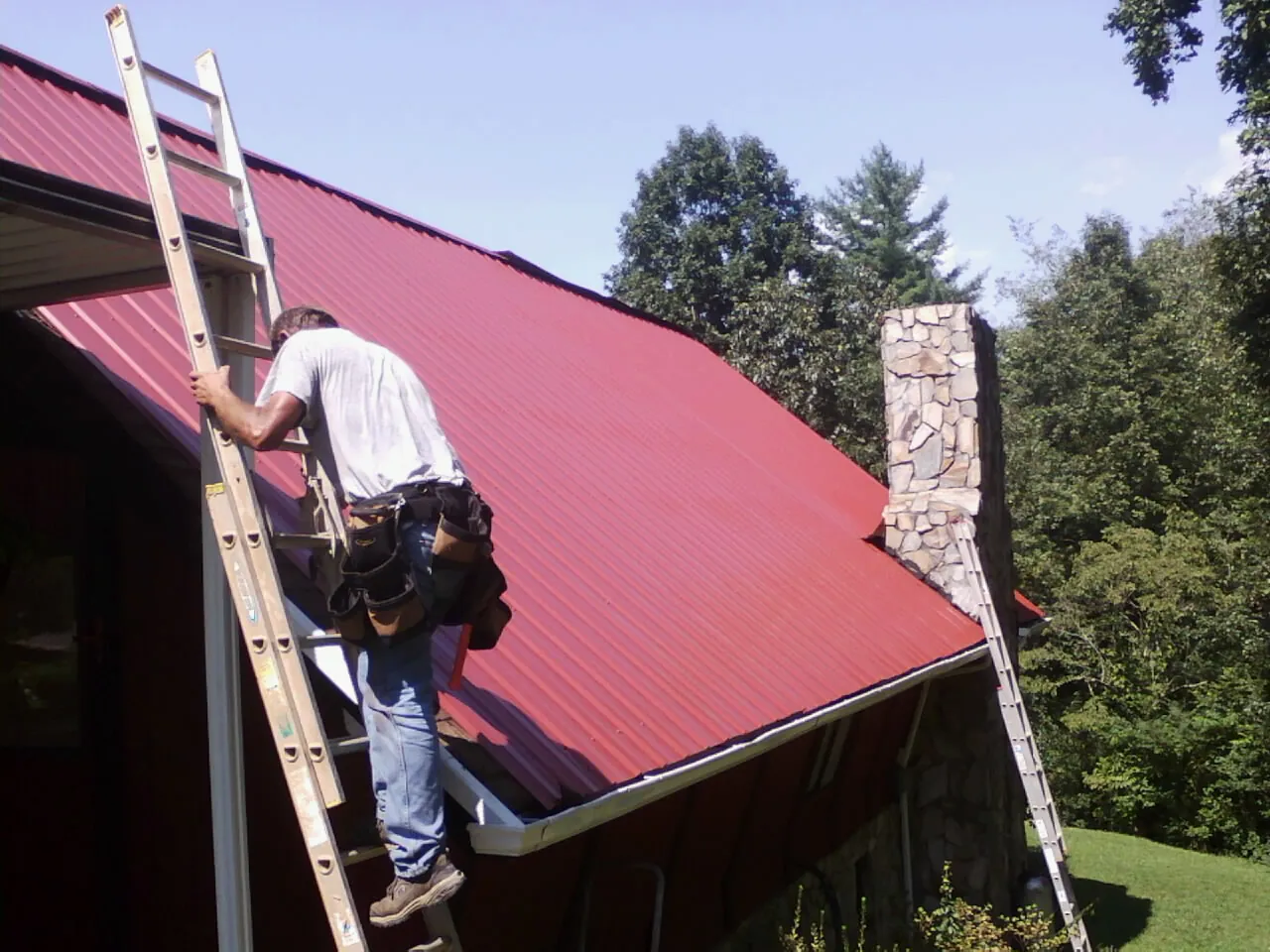 Roofer on a ladder beside a red metal roof and stone chimney — Soffit & Fascia Repair by Roofing Brothers in Toutle
